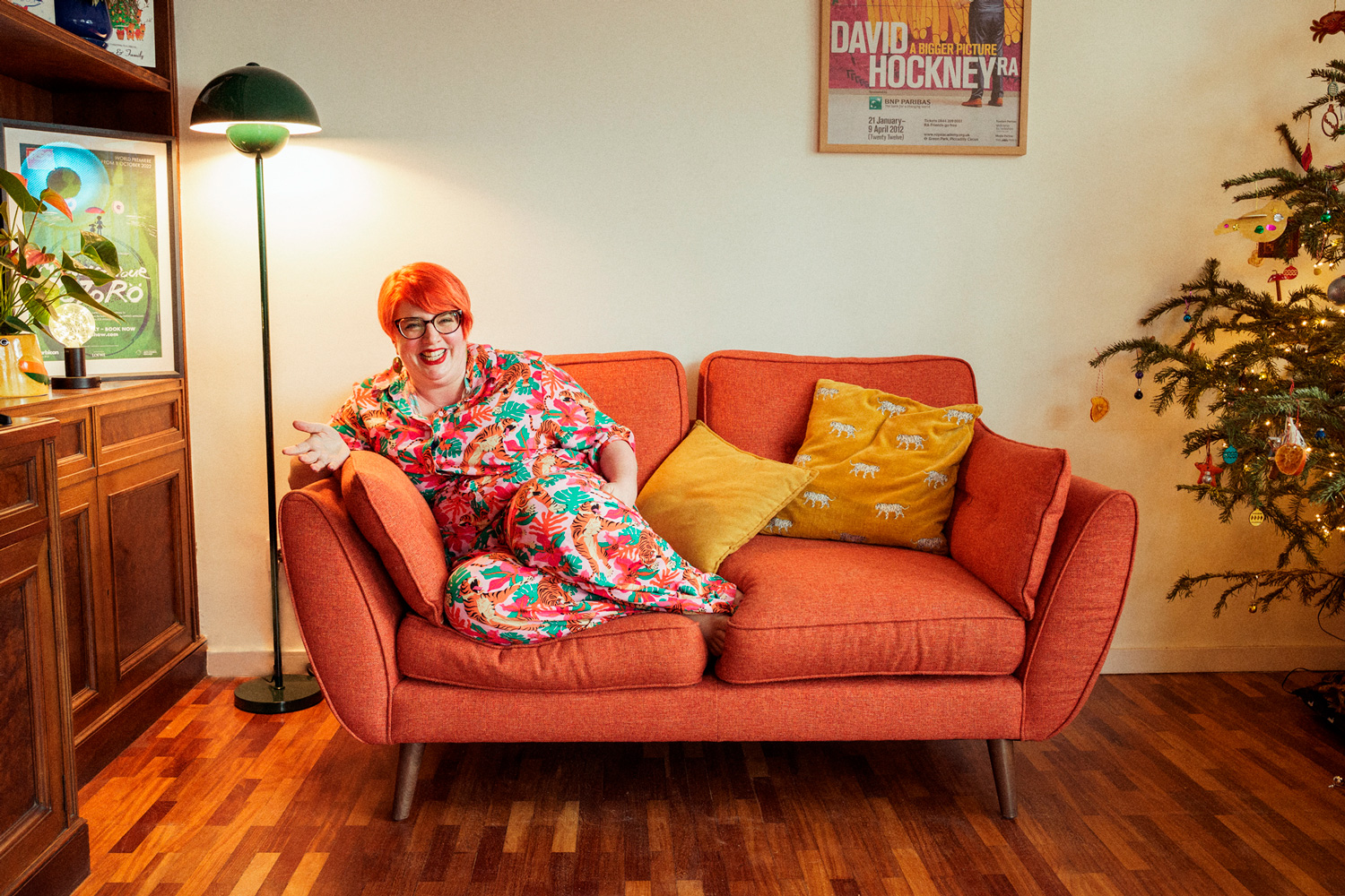 Portrait of Beth sitting on an orange sofa in colorful patterned pajamas, smiling and gesturing, with a Christmas tree beside her